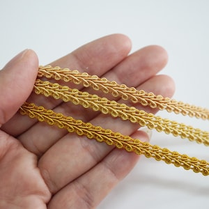 May include: Close-up of three strands of gold-colored decorative trim. The trim has a repeating, ornate design. The trim is held in a hand against a white background. The trim is likely used for sewing, crafting, or embellishing.