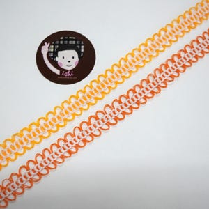 Puede incluir: Dos cintas decorativas con borde festoneado en naranja y amarillo, con centro blanco. Las cintas están en ángulo sobre un fondo blanco. Un logotipo circular con una cara de dibujos animados y la palabra "ichi" está en la esquina superior izquierda.