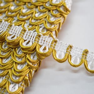 May include: Close-up of decorative trim with a repeating pattern of gold and white scalloped edges. The trim has a woven white base with gold detailing, creating a textured and ornate appearance. Ideal for embellishing garments or home decor projects.