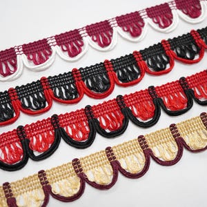 May include: Close-up of several rows of decorative trim in various colours. The trim features a scalloped edge with fringe. Colours include burgundy, white, black, red, and gold. The trim is likely used for sewing or crafting projects.