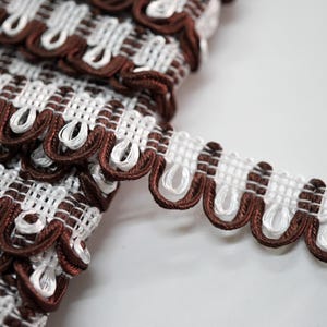 May include: Close-up of decorative trim with a white woven base and brown scalloped edges. The trim has a repeating pattern of brown and white loops, ideal for craft projects.