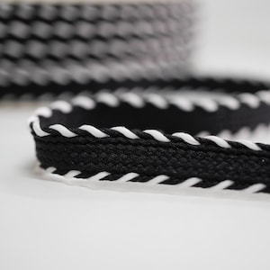 May include: Black and white decorative trim. The trim is black with a white, wavy edge. A roll of the trim is in the background, with a length of the trim in the foreground. The trim is on a white background.