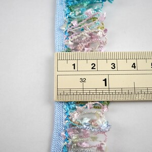 May include: A light blue trim with a multi-colored fringe. The fringe is made of a variety of colors, including pink, green, and blue. The trim is measured with a ruler, showing it is 1 inch wide.