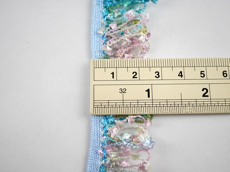 May include: A light blue trim with a multi-colored fringe. The fringe is made of a variety of colors, including pink, green, and blue. The trim is measured with a ruler, showing it is 1 inch wide.