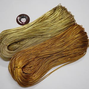 May include: Two bundles of metallic gold cord. One is a lighter, yellowish gold, and the other is a deeper, bronze-toned gold. The cords are tightly wound and appear to be made of a thin, flexible material, suitable for crafts or jewellery making.