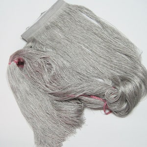 May include: A length of silver-grey fringe, suitable for crafts or decoration. The fringe is closely packed, with long, flowing strands. A pink thread is visible, possibly for binding or accent. The material appears to be soft and pliable.