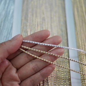May include: Three strands of metallic cord, one silver, one gold, and one with a gold and silver twist, are held in a hand. The cords are all braided and have a shiny finish.