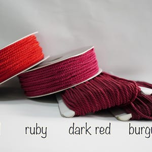 May include: Spools of red, ruby, dark red, and burgundy colored cord. The cord is tightly wound on white spools. The colors are labeled with the corresponding color names in a handwritten font.