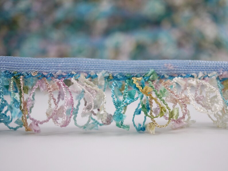 May include: A close-up of a light blue trim with a fringe of colorful threads in shades of pink, blue, green, and gold.