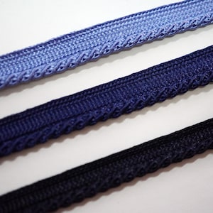 May include: Close-up of three decorative ribbons in shades of blue and black. The ribbons have a braided edge and a textured surface. The colours range from light blue to navy and black, suitable for crafts and sewing projects.