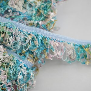 May include: A close-up of a light blue trim with a multi-colored fringe. The fringe is made of thin, curled strands of fabric in shades of blue, green, pink, and white.