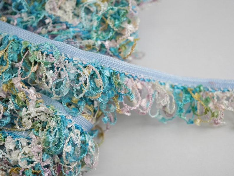 May include: A close-up of a light blue trim with a multi-colored fringe. The fringe is made of thin, curled strands of fabric in shades of blue, green, pink, and white.