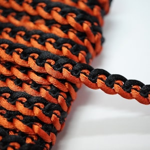 Puede incluir: Borde trenzado negro y naranja con un patrón tejido. El borde está hecho de dos cordones de diferentes colores que están tejidos juntos.