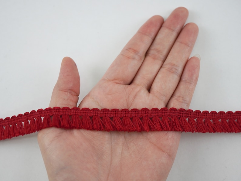 May include: Red tassel trim with a woven base and fringe. The trim is shown on a hand.