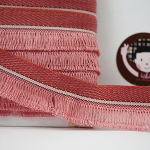 May include: Pink and red striped trim with a fringe. The trim has a woven texture and is approximately 1 inch wide.