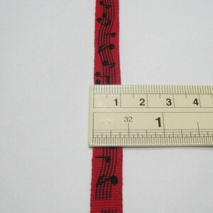 May include: Red ribbon with black musical notes printed on it. The ribbon is measured with a ruler, showing it is 1 inch wide.