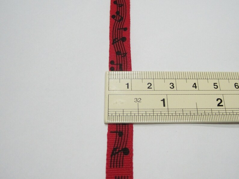 May include: Red ribbon with black musical notes printed on it. The ribbon is measured with a ruler, showing it is 1 inch wide.