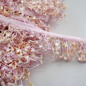 Puede incluir: Borde de flecos rosa y beige con una textura suave y esponjosa. El borde tiene una base tejida y un fleco largo y fluido.