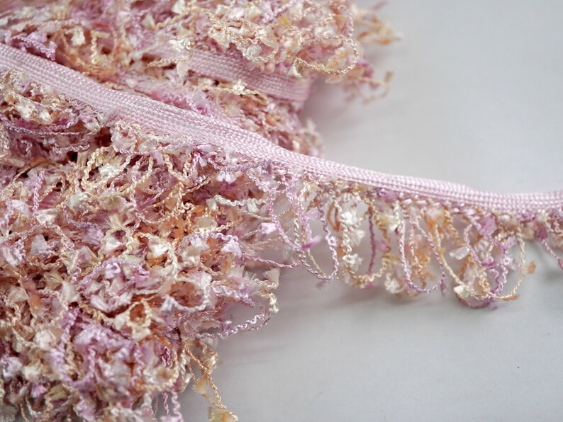 Puede incluir: Borde de flecos rosa y beige con una textura suave y esponjosa. El borde tiene una base tejida y un fleco largo y fluido.