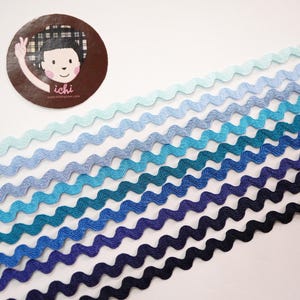 Può includere: Una collezione di nastri decorativi ondulati in tonalità di blu, dall'azzurro chiaro al blu navy scuro. I nastri sono disposti in diagonale, mostrando una sfumatura di colori. Un adesivo circolare con un personaggio dei cartoni animati è in alto a sinistra.