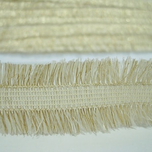 May include: A close-up of a white and gold fringe trim with a woven center. The fringe is soft and fluffy, and the trim is perfect for adding a touch of elegance to any project.