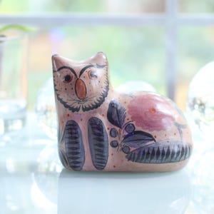 May include: A hand-painted ceramic cat figurine in shades of tan, pink, and black. The cat has an owl-like face with large eyes and a beak-like nose. Floral and leaf patterns decorate the body. The figurine is approximately 6 inches long.