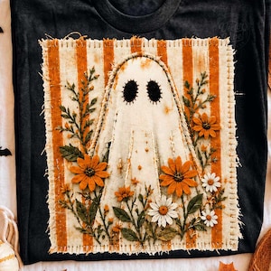 May include: A black t-shirt featuring a rectangular patch with a frayed edge. The patch has orange and white vertical stripes and displays a central white ghost figure with black eyes, surrounded by embroidered orange and white flowers.