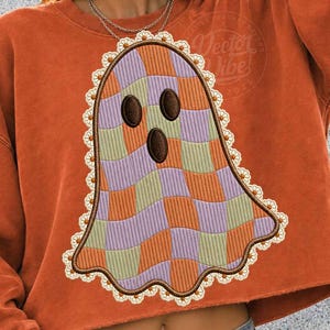 Könnte beinhalten: Ein brennend orangefarbenes bauchfreies Sweatshirt mit einer großen gestickten Geistergrafik. Der Geist hat ein wellenförmiges Karomuster in Lavendel, Salbeigrün und Orange, mit drei dunkelbraunen ovalen Augen und Mund, umrandet von weißer Spitzenborte.