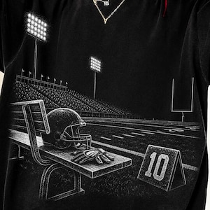 May include: A black t-shirt featuring a white graphic print of a football stadium at night. The design shows a bench with a football helmet and gloves, a yard marker with the number 10, stadium lights, and goalposts on a football field.