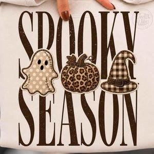 Op de afbeelding: Een crèmekleurig sweatshirt met de donkerbruine tekst SPOOKY SEASON. Het ontwerp bevat een beige geest met stippen, een pompoen met luipaardprint en een heksenhoed met bruin en crème ruitjespatroon, horizontaal gerangschikt in het midden.