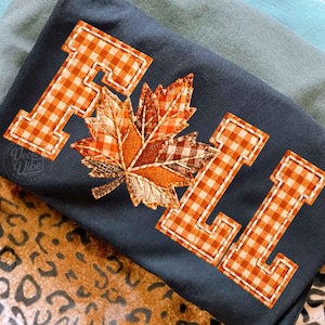 Könnte beinhalten: Ein dunkelgraues T-Shirt mit dem Wort FALL in orange-weiß karierten Blockbuchstaben und einer zentralen Ahornblatt-Applikation aus verschiedenen strukturierten Stoffen in Orange-, Braun- und Beigetönen, die auf den Stoff aufgenäht ist.