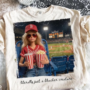 Puede incluir: Camiseta color crema con un gráfico de una muñeca en un estadio de béisbol, sosteniendo palomitas de maíz y usando una gorra y camiseta de béisbol rojas. El texto "literally just a bleacher creature" está impreso debajo.