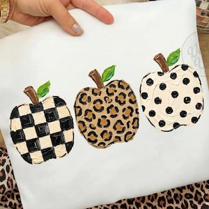 May include: White decorative pillow featuring three painted pumpkins. One pumpkin has a black and white checkerboard pattern, another has a leopard print, and the third has black polka dots. Each pumpkin has a brown stem and green leaf.