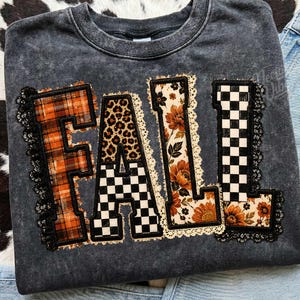 May include: A charcoal gray acid-wash crewneck sweatshirt featuring the word FALL in large block letters. Each letter has a unique pattern: orange plaid, leopard print, floral print, and black-and-white checkerboard, all outlined with black lace.