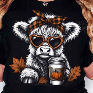 Può includere: Una t-shirt nera con la grafica di una soffice mucca highland bianca che indossa occhiali da sole a forma di cuore e una fascia a quadri. La mucca tiene in mano una tazza di caffè con un'illustrazione di zucca, circondata da foglie autunnali.