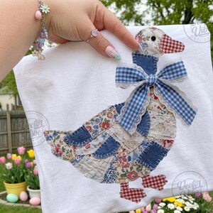 May include: White t-shirt featuring a patchwork goose design. The goose is made of floral and denim fabrics, with a blue gingham bow and lace trim. The goose's beak and feet are red and white checkered fabric. The design is centered on the shirt.