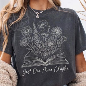 May include: Dark gray t-shirt featuring a floral design emerging from an open book. The text "Just One More Chapter" is written below the book. The shirt has a vintage, worn look.