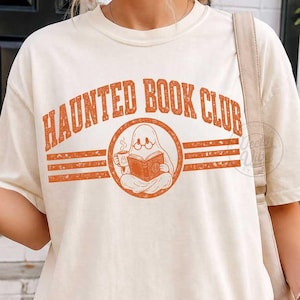 Puede incluir: Una camiseta de manga corta color crema con el texto naranja "HAUNTED BOOK CLUB" en fuente arqueada. El gr&aacute;fico incluye un emblema circular central de un fantasma con gafas que lee un libro mientras sostiene una taza de caf&eacute;.