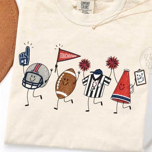 May include: A cream-colored t-shirt featuring a graphic of four anthropomorphic football-themed characters: a helmet with a foam finger, a football holding a red pennant labeled TOUCHDOWN, a referee, and a megaphone holding a play clipboard.