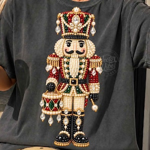 May include: A charcoal grey t-shirt featuring a large graphic of a nutcracker soldier adorned with faux pearls, red and green beads, and clear crystals. The nutcracker wears a tall hat, holds a drum, and has a detailed moustache.