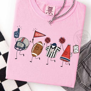 May include: A light pink t-shirt featuring a graphic of cartoon football characters including a helmet with a number one foam finger, a football with a touchdown flag, a referee shirt with pom-poms, and a megaphone holding a play clipboard.