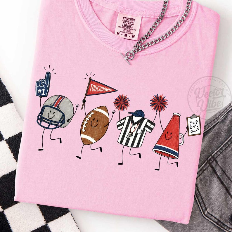 May include: A light pink t-shirt featuring a graphic of cartoon football characters including a helmet with a number one foam finger, a football with a touchdown flag, a referee shirt with pom-poms, and a megaphone holding a play clipboard.
