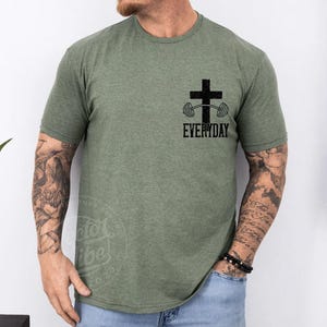 Puede incluir: Camiseta verde oliva con un gr&aacute;fico negro de una cruz y una barra con pesas, con la palabra "EVERYDAY" debajo. La camiseta tiene mangas cortas y cuello redondo. La persona lleva vaqueros azules y una pulsera de cuentas negras.
