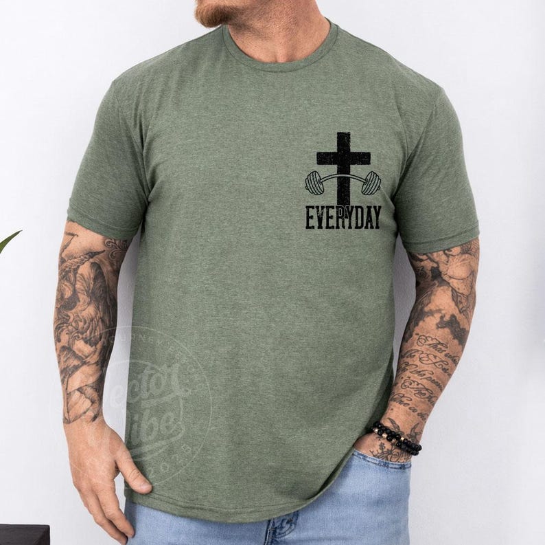 Puede incluir: Camiseta verde oliva con un gr&aacute;fico negro de una cruz y una barra con pesas, con la palabra "EVERYDAY" debajo. La camiseta tiene mangas cortas y cuello redondo. La persona lleva vaqueros azules y una pulsera de cuentas negras.