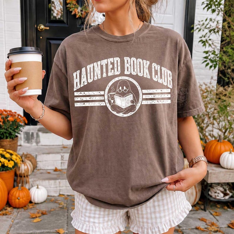 Puede incluir: Una camiseta marr&oacute;n de manga corta con el texto blanco Haunted Book Club en una fuente arqueada sobre un gr&aacute;fico circular de un fantasma con gafas leyendo un libro y sosteniendo una taza, con l&iacute;neas horizontales flanqueando el dise&ntilde;o.