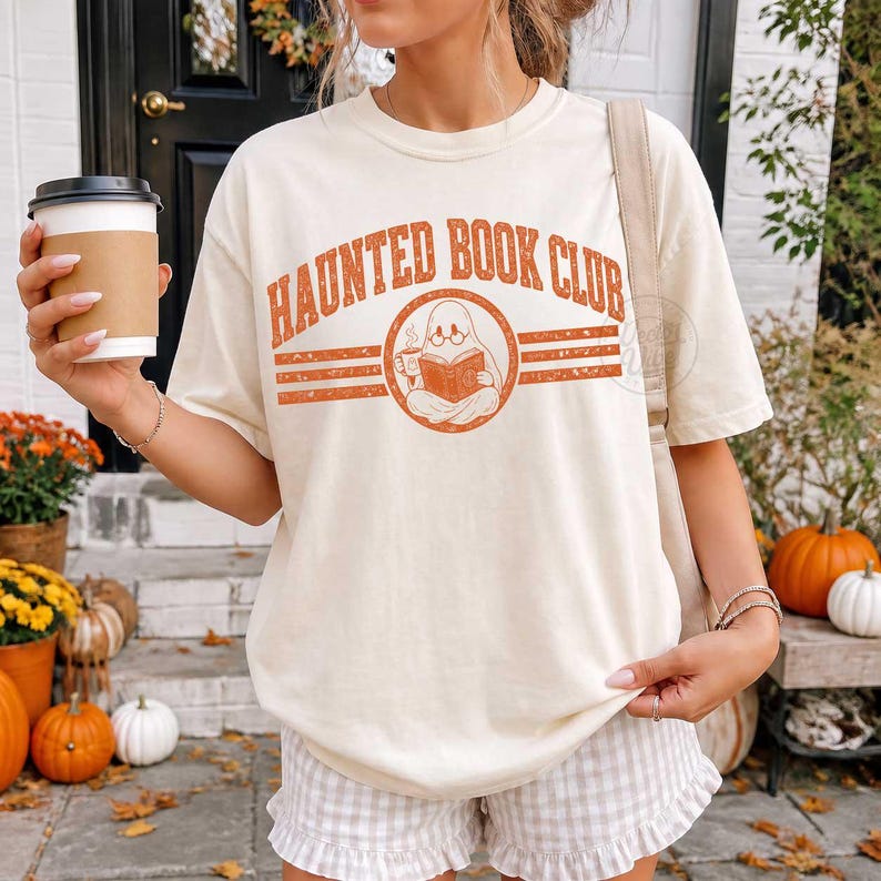 Puede incluir: Una camiseta de manga corta color crema con el texto naranja "HAUNTED BOOK CLUB" en fuente arqueada. El gr&aacute;fico incluye un emblema circular central de un fantasma con gafas que lee un libro mientras sostiene una taza de caf&eacute;.