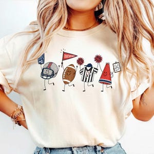 May include: A cream-colored t-shirt featuring a graphic print of four cartoon characters representing American football: a helmet with a foam finger, a football holding a red pennant labeled TOUCHDOWN, a referee, and a megaphone with a play sheet.