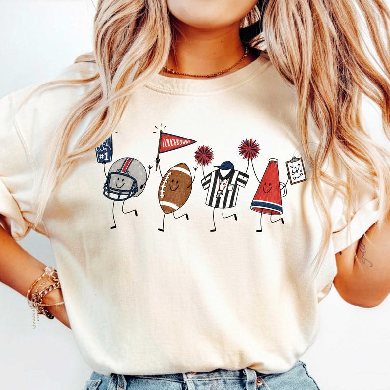 May include: A cream-colored t-shirt featuring a graphic print of four cartoon characters representing American football: a helmet with a foam finger, a football holding a red pennant labeled TOUCHDOWN, a referee, and a megaphone with a play sheet.