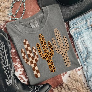 May include: A grey t-shirt with three cactus designs: a brown and white chequered cactus, a leopard print cactus, and a dotted cactus. The shirt is styled with denim jeans, a cowboy hat, and boots.