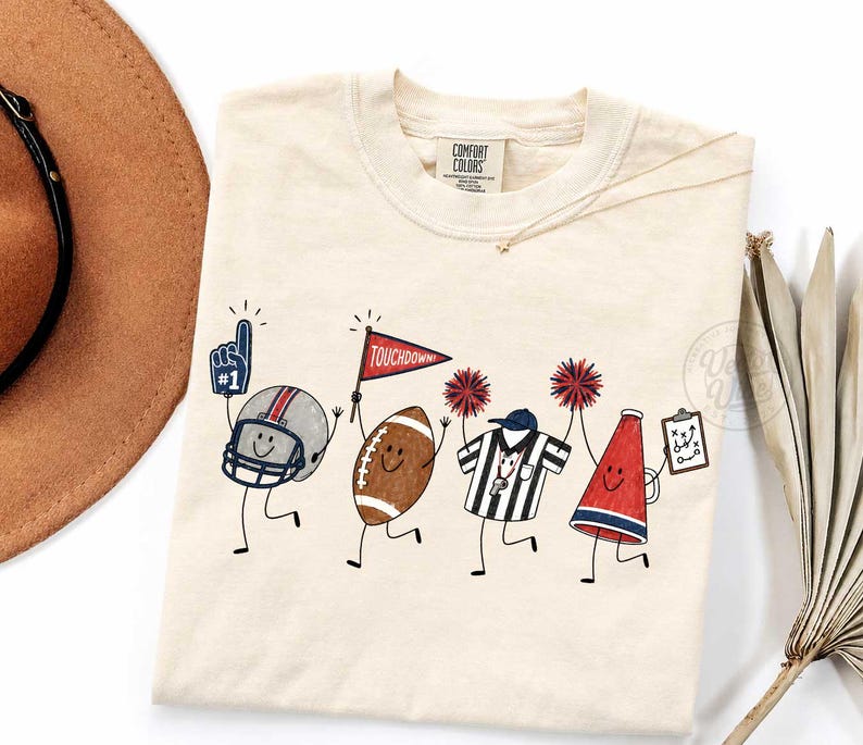 May include: A cream-colored t-shirt featuring a graphic of four anthropomorphic football-themed characters: a helmet with a foam finger, a football holding a red pennant labeled TOUCHDOWN, a referee, and a megaphone holding a play clipboard.